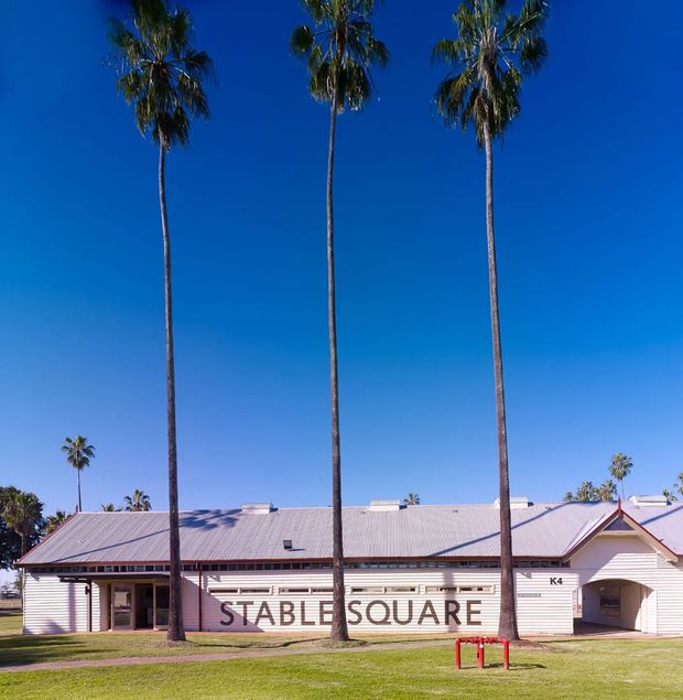 Exterior photograph of a low-rise commercial or community building labelled “STABLE SQUARE K4”. The building has a long, pale weatherboard facade, a broad low-pitched corrugated metal roof, simple gabled end detailing, and small rectangular windows and doors. Several tall palm trees stand prominently in front, with a manicured lawn and vivid blue sky. The image has a bright, warm, resort-like outdoor character.