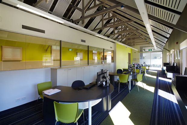 A bright contemporary open-plan commercial office or educational workspace with shared meeting tables, computer workstations, and lime-green and dark-blue chairs. A long glass-partitioned room with pale cabinetry runs along one wall. The space has dark striped carpet, lime-green accent walls, extensive glazing, and a distinctive exposed high ceiling with timber-toned structural trusses and slatted acoustic panels.