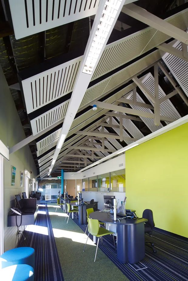 A bright contemporary commercial interior, likely a café, lounge, or informal workplace, with rows of small round tables, mixed chairs, upholstered lounge seating, and a long service or counter area. The space has a dramatic exposed pitched roof structure with painted steel trusses, white slatted acoustic ceiling panels, linear lighting, and a vivid lime-green feature wall. Dark striped carpet, glass partitions, and daylight create a high-ceilinged, open atmosphere.
