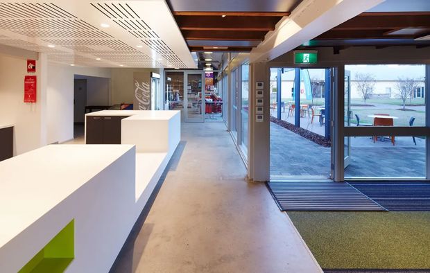 A bright, contemporary commercial interior foyer or reception area with white geometric built-in counters, a polished concrete floor, exposed dark timber ceiling beams, patterned acoustic ceiling panels, and extensive glazing. The space opens to an outdoor paved area through glazed doors, with an illuminated emergency exit sign and a glimpse of a refreshment or retail area beyond.