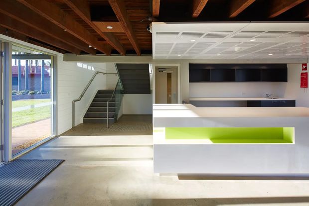 A bright contemporary commercial reception and entry area with a white reception desk featuring a vivid lime-green inset, dark built-in cabinetry behind it, an exposed timber-beam ceiling, polished concrete flooring, large glazed doors, and a stairway leading to an upper level. The space has an industrial-institutional character with strong natural light and clean, functional finishes.
