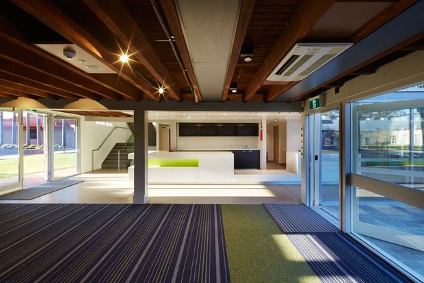 A bright contemporary commercial interior with an open reception or communal workspace. The space features exposed dark timber ceiling beams, a large central white built-in counter with a lime-green accent panel, dark blue cabinetry, extensive glazing and glazed entry doors, a carpeted floor, and a stairway leading to an upper level. Strong daylight enters from the windows and doors.