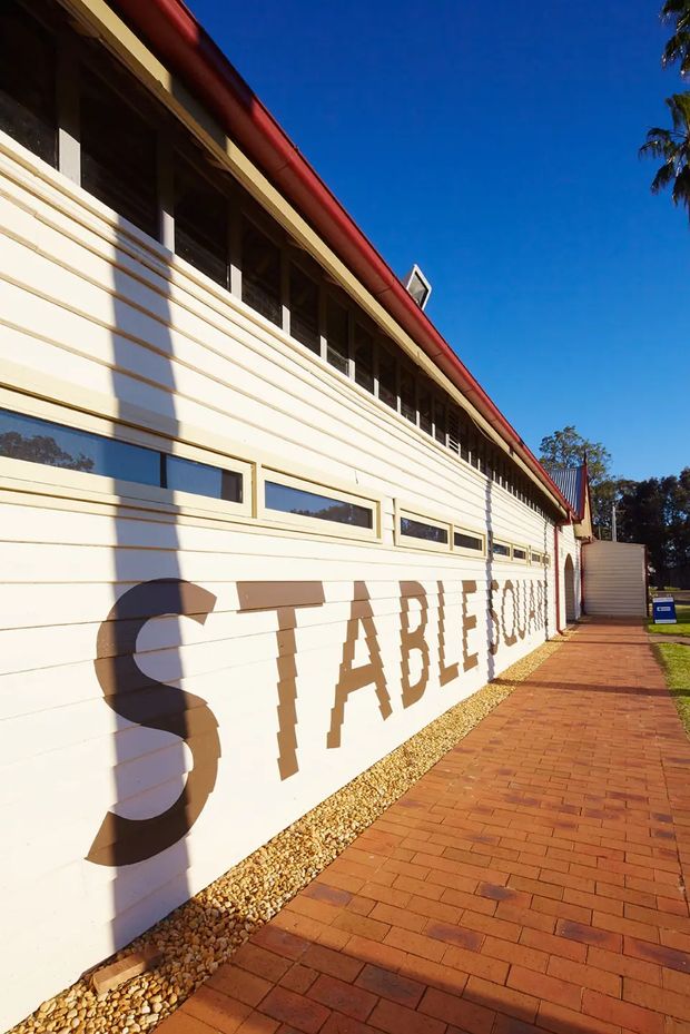Exterior view of a long, single-storey cream weatherboard building with a red pitched roof, high clerestory-style windows, and large painted lettering on the wall. A red brick-paved walkway runs alongside the building, with gravel edging and trees visible beyond. The image is strongly sunlit with deep shadows and a clear blue sky.