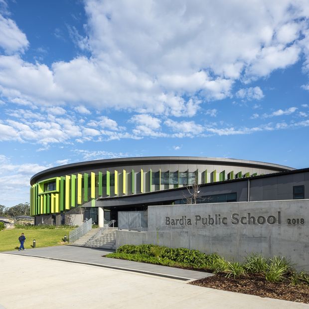 Exterior view of Bardia Public School, a contemporary institutional building with a sweeping curved roofline, dark cladding, extensive glazing, and a vivid vertical lime-green and yellow sun-shading facade. Concrete entry signage, stairs, landscaping, and a broad paved forecourt are visible under a bright partly cloudy sky.