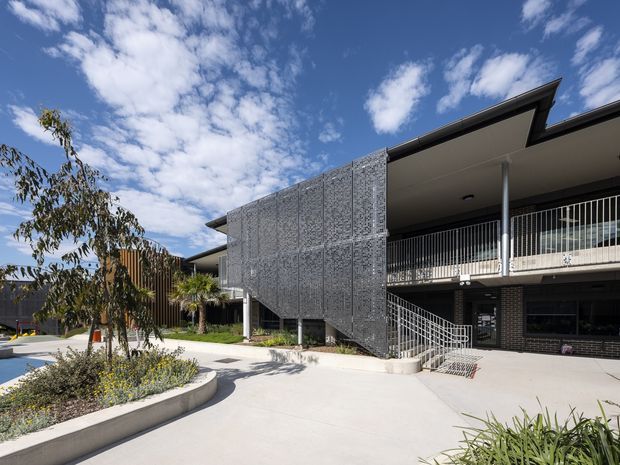 Exterior view of a contemporary multi-storey building with a large perforated metal screening feature, dark brickwork, slim metal balcony railings, and a broad concrete-paved courtyard. Landscaped garden beds and palm plantings sit in the foreground beneath a bright blue sky.