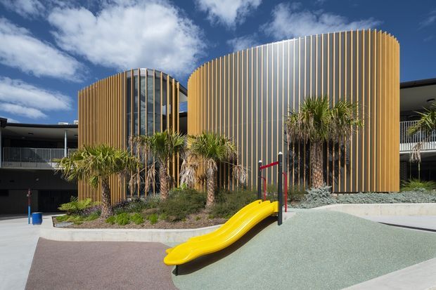 Exterior view of a contemporary multi-level building with two prominent rounded volumes wrapped in vertical timber-toned screening and glazing. A landscaped play area with palm-like planting, a bright yellow slide, safety surfacing, and handrails occupies the foreground under a partly cloudy blue sky.