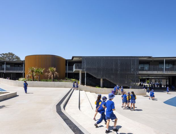 Wide exterior view of a contemporary multi-storey school or institutional building surrounding a broad paved courtyard. The building features dark horizontal glazing and metal elements, a curved vertical timber-look screen on the left, and a large dark perforated or mesh facade panel over a central stair zone. Students in blue uniforms occupy the open concrete forecourt, with integrated linear drainage channels, stepped access, planting beds, and low retaining walls.