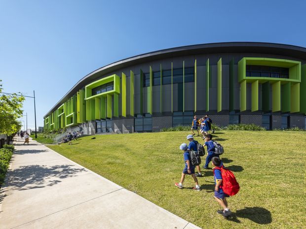 Exterior view of a large contemporary civic or educational building with a sweeping curved roofline, dark glazed facade, and vivid lime-green vertical fins and projecting window frames. A gently sloping lawn and concrete pedestrian path lead toward the building, with school-aged children visible outside. The composition emphasizes the building's bold facade and landscaped approach under a clear blue sky.