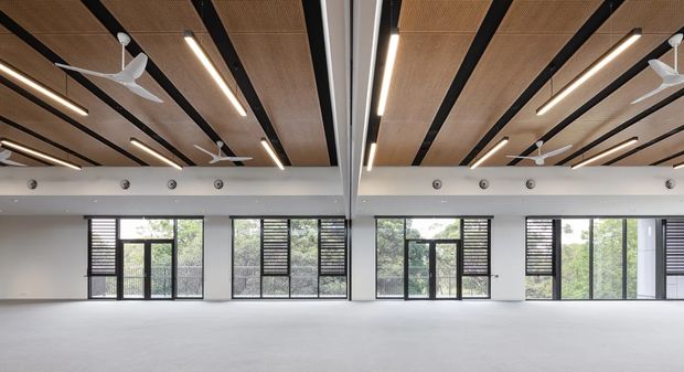 A large, open-plan contemporary interior with a broad expanse of light flooring, white walls, extensive floor-to-ceiling glazing, and black-framed doors and windows fitted with horizontal blinds. The ceiling features warm timber acoustic panels arranged between exposed dark structural beams, linear strip lighting, and multiple white ceiling fans. The space appears bright, spacious, and suitable for a commercial, community, or multipurpose use.