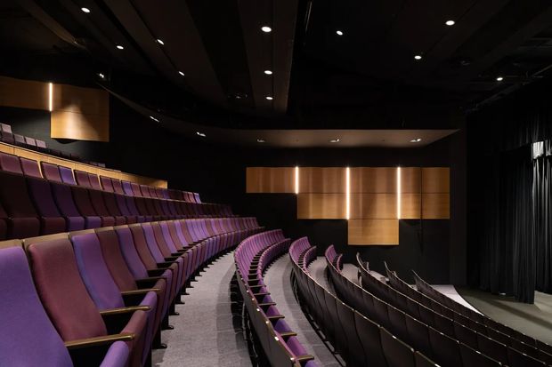 Interior view of a modern auditorium or theatre with steeply tiered, curved rows of upholstered purple and burgundy seats. Dark acoustic walls and ceiling surround a stage area with black curtains, while warm timber or wood-toned illuminated wall panels form a geometric feature at the front. Recessed ceiling lighting creates a dramatic, refined atmosphere.