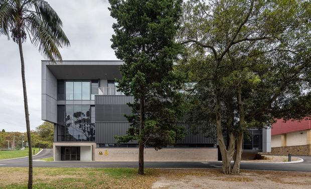 A contemporary multi-storey institutional or commercial building viewed from across a landscaped frontage. The facade combines dark vertical metal cladding, pale concrete or rendered panels, extensive floor-to-ceiling glazing, and a broad flat projecting roof canopy. Mature trees and a palm partially screen the building, while a paved driveway and accessible entrance are visible at ground level.