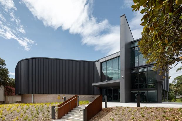 Exterior view of a contemporary multi-level building with a strongly sculptural form. The facade combines a dark vertically ribbed curved wall, extensive blue-grey glazing, and a tall charcoal blade-like structural element. A concrete entry stair with warm timber-toned side walls leads to the entrance, surrounded by landscaped planting and mature trees.