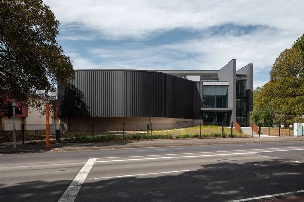 Street-facing view of a contemporary multi-storey building with a strong geometric composition. The facade combines a large dark vertically ribbed cladding volume with pale rendered or concrete sections, extensive glazing, projecting angular forms, and a recessed upper-level terrace or balcony. The building sits behind a low fence and landscaped verge along a marked urban road, with mature trees framing the scene.