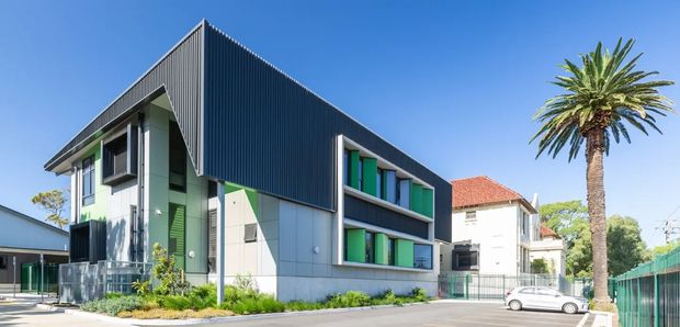 Exterior view of a contemporary multi-storey building with a sharply angled corrugated dark metal upper facade, pale panel cladding, projecting window bays with bright green and turquoise accents, and a landscaped frontage. A palm tree and neighbouring traditional tiled-roof homes are visible around the site.