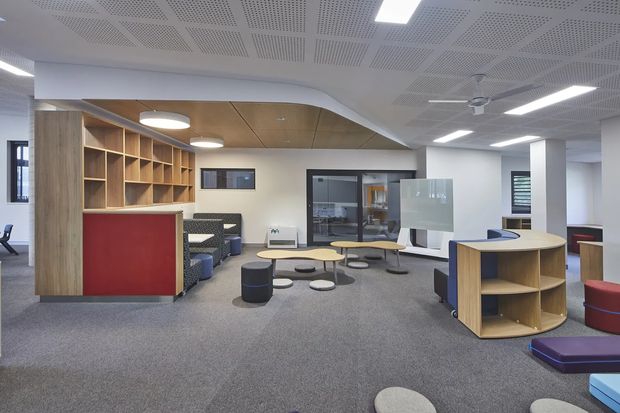 A contemporary open-plan commercial interior, likely a school or workplace breakout and collaboration area. The carpeted room contains modular lounge seating, low round tables, upholstered stools, curved timber shelving and divider joinery, and a red-accented built-in shelving unit. White walls, acoustic perforated ceiling panels, linear LED lighting, and timber ceiling panels create a bright, functional environment.
