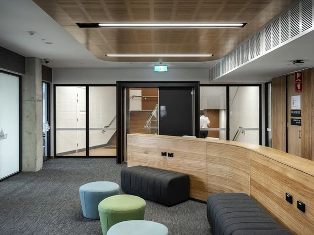 Contemporary commercial reception or waiting area with a curved timber-clad reception desk, upholstered modular stools, dark carpet, glazed internal partitions, a visible stairwell, and a linear timber ceiling with recessed strip lighting. Black-framed glass doors and partitions, exposed concrete, and ventilation grilles contribute to a refined office fit-out.