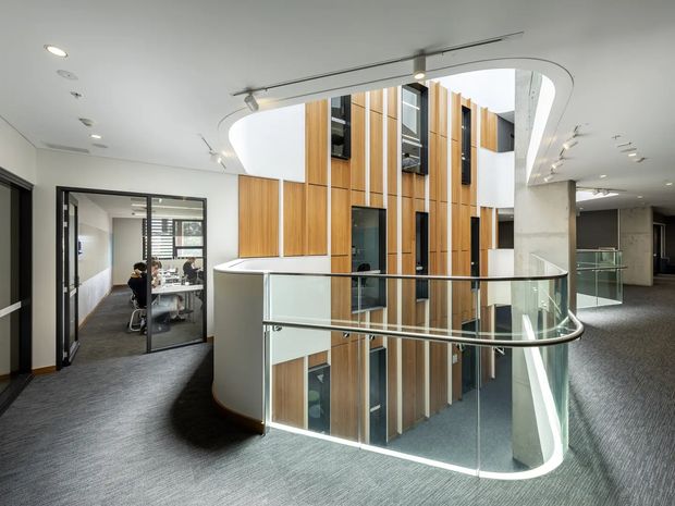 A bright contemporary commercial office interior viewed from an upper-level landing, centred on a curved glass balustrade overlooking a multi-storey internal atrium. The atrium features tall vertical timber wall panels, white curved ceiling forms, and visible windows and circulation spaces. A glazed meeting room with desks and occupants is visible to the left, while carpeted corridors extend to the right and rear.