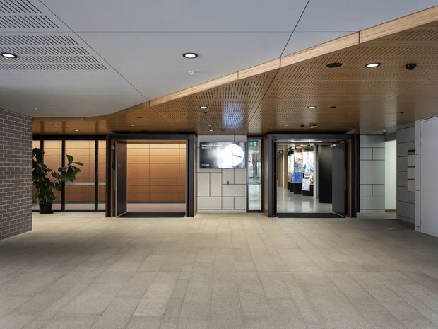 A modern commercial building lobby or lift foyer with a spacious tiled floor, timber slatted acoustic ceiling and bulkhead, recessed lighting, dark-framed glazed lift entrances, light tiled wall panels, and a brick feature wall. Digital signage and a glimpse of a corridor or retail area are visible beyond the doors.