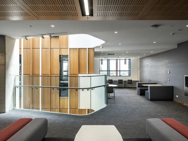 A spacious contemporary commercial interior, likely an office or shared lounge, viewed from an upper-level landing around a curved glazed balustrade. The space features grey carpet, a grey tiled feature wall, timber-lined vertical partitions, exposed concrete columns, soft seating, meeting tables, large windows, recessed lighting, and a slatted timber acoustic ceiling with a curved ceiling opening.