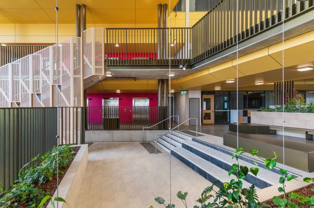 A contemporary multi-level interior atrium or communal entrance space with a warm ochre ceiling and soffits, dark vertical metal balustrades, exposed structural columns, suspended lighting, and broad tiled stairs. Raised planter beds with greenery define the circulation areas, while bright magenta doors and wall panels provide a strong accent. The space includes upper-level walkways and extensive internal glazing, suggesting a commercial, institutional, or apartment-building common area.