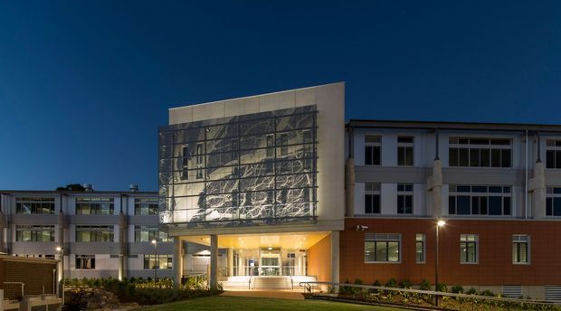Night-time exterior view of a large institutional or commercial building complex. The composition is dominated by a projecting entrance pavilion with a tall illuminated patterned glass or metal feature screen, canopy, and glazed lobby. The adjoining multi-storey wings combine reddish-brown masonry panels, pale rendered or concrete elements, horizontal window bands, and a covered accessible entrance walkway. Landscaped planting and a sloped access path are visible in the foreground against a deep blue sky.