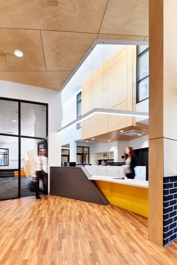 Bright contemporary commercial interior, likely a reception or communal workspace, featuring an open double-height volume, warm plywood ceiling and wall panels, black-framed internal glazing, light timber flooring, and a white built-in counter with a vivid yellow base. A dark angular reception desk and black brick-clad column add contrast, while a suspended linear light defines the upper workspace.