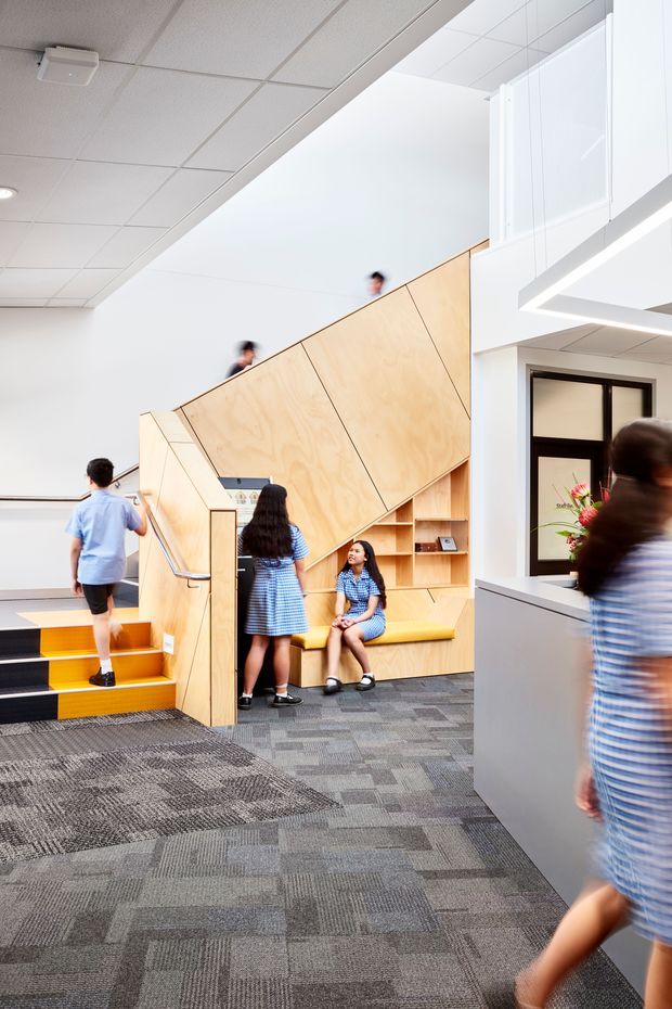 Bright contemporary school or institutional interior featuring a sculptural plywood stair and mezzanine structure, integrated timber seating and shelving, grey carpet tiles, and white acoustic ceiling panels. Several students in uniforms use the stair and seating area, while a reception or service counter is visible at right.