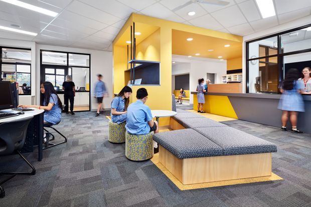 Bright contemporary commercial interior, likely a reception and collaborative breakout area. The open-plan space features a grey patterned carpet, white acoustic ceiling tiles, black-framed glazed partitions, a vivid yellow architectural feature wall and ceiling recess, dark grey reception counter, timber-clad built-in seating, and patterned upholstered benches and stools. People are using desks and informal seating areas, with additional workspaces visible through the glazing.