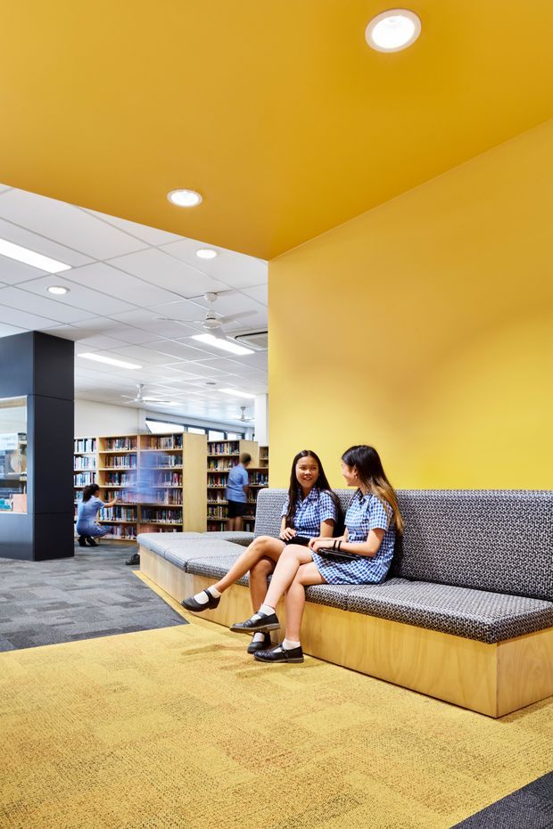 Bright contemporary library or learning commons with a bold mustard-yellow ceiling and wall, recessed downlights, and a large built-in timber banquette upholstered in patterned grey fabric. Two students sit on the seating while bookshelves, study areas, ceiling fans, and fluorescent ceiling panels extend into the background. The floor combines grey carpet tiles with a large yellow carpeted area.