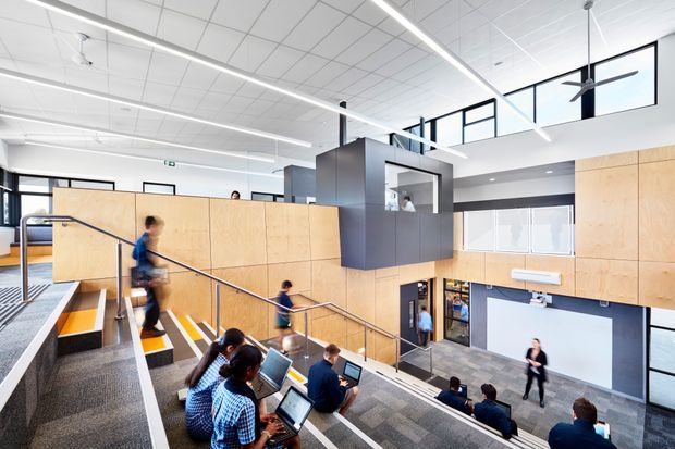 A bright contemporary multi-level commercial interior resembling a lecture theatre, studio, or educational workspace. Tiered carpeted seating and stairs descend toward an open lower-floor area, with people working on laptops and walking through the space. Pale timber wall panelling, dark grey built-in volumes, extensive internal glazing, exposed suspended ceiling tiles, linear strip lighting, and large ceiling fans define the architecture. The space has a spacious, open atrium-like character with strong visual connections between levels.