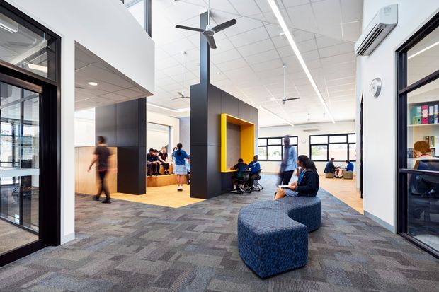 Bright contemporary commercial interior, likely an educational or workplace common area, with an open-plan circulation and informal seating zone. The space features a patterned carpet, white ceiling with linear lighting and ceiling fans, black architectural partitions, a vivid yellow recessed niche, extensive black-framed glazing, and people using lounge and work areas. Offices or rooms open from the central space on both sides.