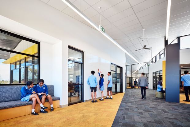 Bright contemporary institutional or commercial interior showing a wide corridor and open circulation area. The space has white walls, a suspended acoustic tile ceiling, linear strip lighting, large internal glazed partitions, grey patterned carpet, warm yellow-orange flooring, and built-in upholstered bench seating. Several people are present, with open work or communal spaces visible through glazing. The corridor appears spacious, airy, and highly functional.