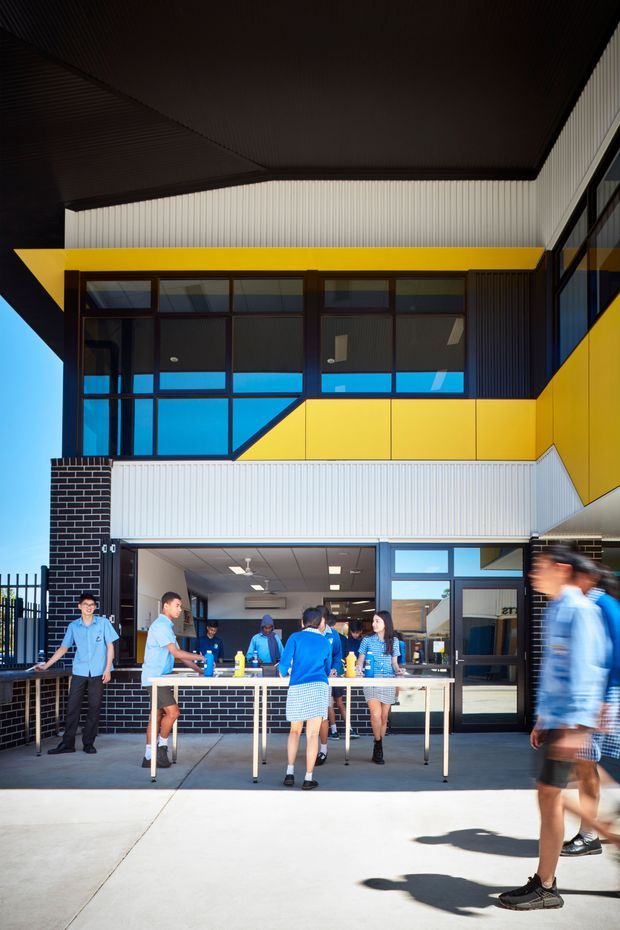 Exterior view of a contemporary multi-storey institutional or commercial building with a covered entry terrace. The facade combines black brick, white vertically ribbed cladding, dark-framed glazing, and bold yellow geometric panels. People gather around outdoor tables beneath the overhang, while a bright blue sky is visible at the side.