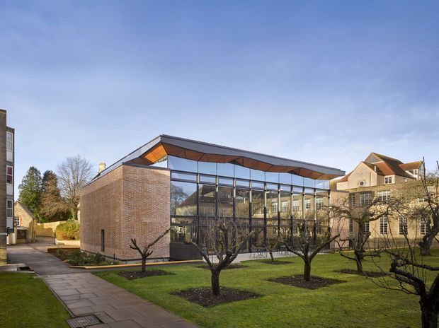 Exterior view of a contemporary institutional or commercial building set within a landscaped courtyard. The building combines a solid warm-toned brick side wall with extensive floor-to-ceiling glazing, dark metal framing, and a distinctive angular roof with exposed warm timber soffit panels. Mature leafless trees, lawns, paved paths, and neighbouring traditional buildings surround the structure.