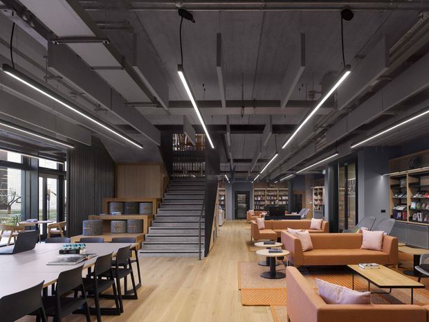 A spacious contemporary commercial interior combining a lounge, library, dining or collaborative workspace, and an open stair. The room features exposed concrete ceilings with visible services, linear suspended lighting, dark ribbed wall panels, warm timber flooring and joinery, built-in bookshelves, and clusters of orange and blush upholstered sofas around low coffee tables. Large windows and glazed doors line the left side, while a broad central stair leads to an upper mezzanine or additional level.