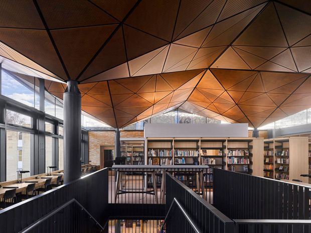 Interior view of a contemporary library or educational reading space, centred on a stair descending between black metal balustrades. Extensive bookshelves, study tables, large windows, and a dramatic faceted timber ceiling with exposed dark structural lines define the space. The warm ceiling contrasts with the black stair and framing elements, creating an expressive, civic interior.