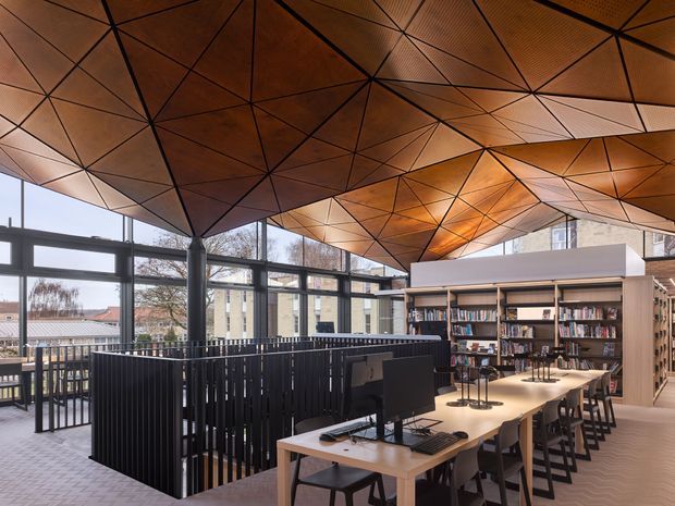 A contemporary library or study area with long shared timber desks, computers, task lighting, shelving filled with books, and black metal railings around an internal void or stair opening. The space is enclosed by extensive dark-framed glazing and features a highly distinctive faceted timber ceiling with geometric black framing and warm integrated lighting. The atmosphere is warm, refined, and institutional yet inviting.
