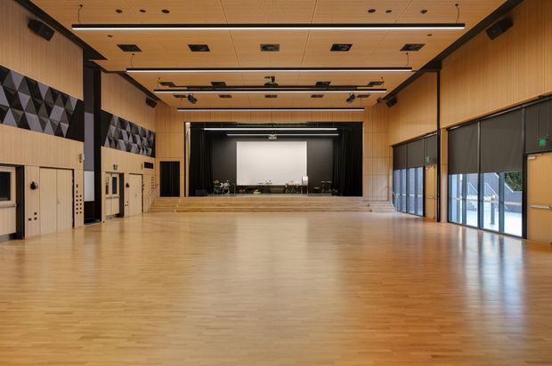 Large modern multipurpose hall or auditorium with a polished timber floor, extensive timber wall and ceiling panelling, acoustic geometric wall panels, exposed black structural framing, integrated lighting and speakers, and a raised stage with projection screen and curtains. Full-height glazed doors and windows line one side, with roller blinds providing light control.