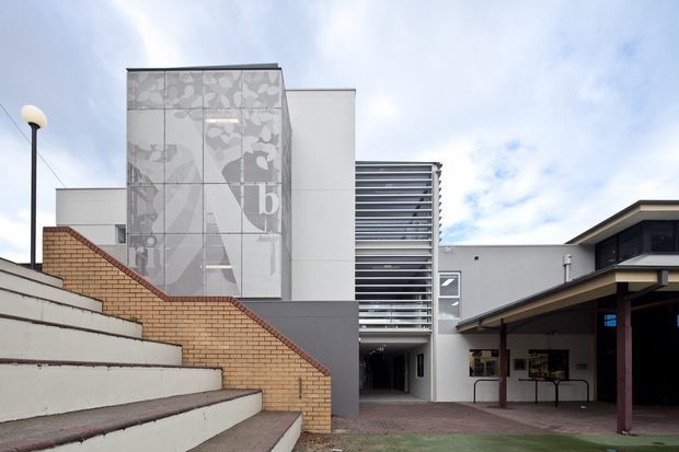 Exterior view of a modern multi-level institutional or commercial building, approached by broad concrete steps. The facade combines white rendered panels, grey wall sections, exposed warm brick stair walls, extensive horizontal metal screening, and a tall translucent graphic panel. A covered entry or walkway is visible to the right, with a paved forecourt and overcast sky.