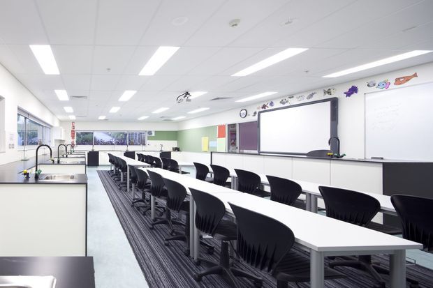 Bright, spacious classroom or teaching laboratory with rows of long white tables and black chairs, whiteboard and large projection screen, extensive windows, pale green walls, and a suspended acoustic tile ceiling with recessed fluorescent lighting. The room has a clean, functional institutional fit-out.