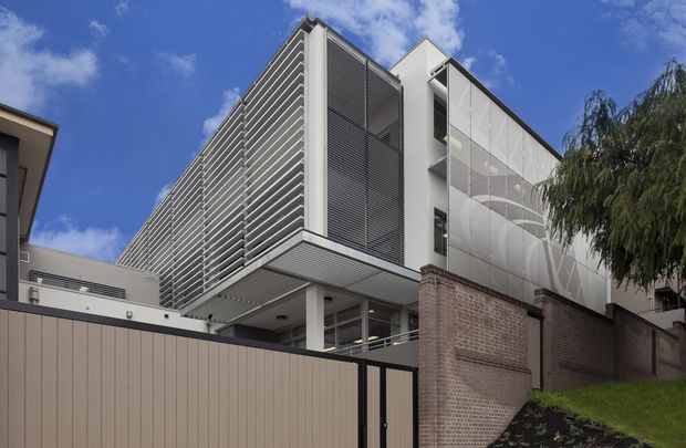 Exterior view of a contemporary multi-storey building on a sloped urban site. The facade combines extensive horizontal metal louvres, smooth white wall panels, glazed openings, and a projecting upper volume supported by slender columns. A brick boundary wall, dark metal gates, retaining elements, and landscaped planting occupy the foreground beneath a bright blue sky.