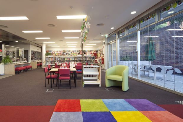 Bright contemporary library or public reading space with rows of bookshelves, study tables and chairs, a service or reception counter, and a large glazed facade overlooking an adjacent courtyard. The interior has dark carpet flooring, white ceiling panels with recessed and linear lighting, colourful shelving and furniture, a sculptural lime-green lounge chair, and a bold multicoloured geometric carpet or floor feature in the foreground.