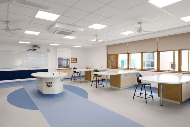 Bright, spacious contemporary commercial learning or collaboration room with white walls, a suspended acoustic tile ceiling, multiple ceiling fans and recessed fluorescent lights, large windows with roller blinds, and several white worktables with stools. A curved dark-blue wall feature, central circular island table, whiteboard, and blue-and-grey patterned flooring create a clean educational or activity-focused environment.