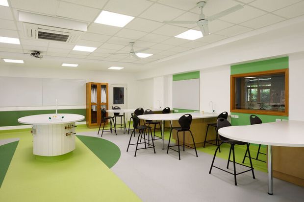 Bright contemporary commercial break room or staff cafeteria with a playful green-and-white interior. The space features several round white tables, black bar stools and chairs, a long communal counter, a curved green-and-white floor pattern, green wall accents, timber-framed interior glazing, a suspended acoustic tile ceiling, fluorescent panel lighting, ceiling fans, and an air-conditioning unit. The layout is open and functional, with a kitchenette or service counter visible at the right.