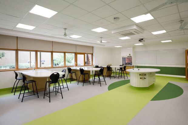 Bright, open commercial learning or activity room with rows of high tables and black stools, a central rounded island counter, extensive timber-framed windows, green-and-lime patterned flooring, and a suspended acoustic tile ceiling with recessed lights and ceiling fans. The space has a clean, functional institutional fit-out.