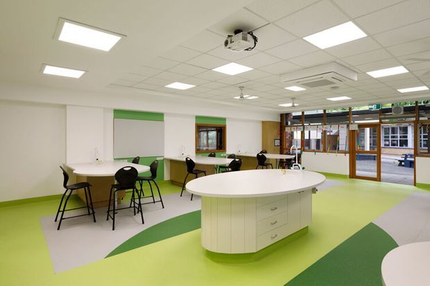 Bright, open-plan commercial interior with a central curved white service island, perimeter work counters, round tables, black chairs, extensive glazing, and a colourful geometric green, lime, and pale grey floor. The space has a suspended acoustic tile ceiling with recessed fluorescent lighting, ceiling fans, and a projector, suggesting a flexible educational or community facility.