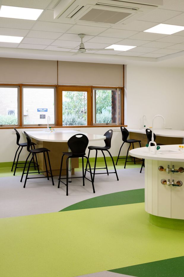 Bright commercial or institutional interior, likely a school or childcare dining and activity area. The room has curved white tables and a curved service island with exposed plumbing fixtures, black stackable chairs and stools, large timber-framed windows, and a suspended acoustic tile ceiling with recessed fluorescent lighting, ceiling cassette air conditioning, and a central fan. A bold green and pale lime resilient floor pattern adds colour to the otherwise neutral space.