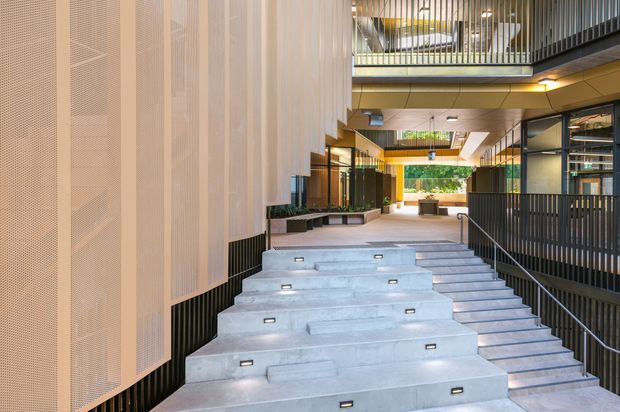 A contemporary multi-level commercial or institutional atrium/courtyard with broad stepped concrete seating or stairs, integrated step lights, black metal balustrades, perforated light-coloured facade panels, extensive glazing, and an upper-level walkway or balcony. The space is open, bright, and strongly defined by modern architectural detailing.