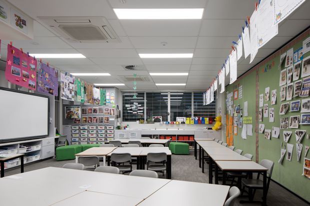 A bright, organised classroom or learning space with rows of grouped tables and chairs, a large whiteboard, extensive student artwork and educational displays, storage shelving, and a green bulletin-board wall. The room has a suspended acoustic ceiling with recessed fluorescent lighting and visible air-conditioning units.