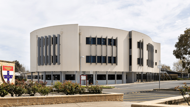 A contemporary multi-storey institutional building with a curved, pale rendered facade and extensive vertical sun-shading fins around dark windows. The structure is elevated on slender columns at ground level, with a broad paved forecourt, landscaped rose beds, signage, and an external stairway. The image is a clear exterior architectural photograph under a bright overcast sky.